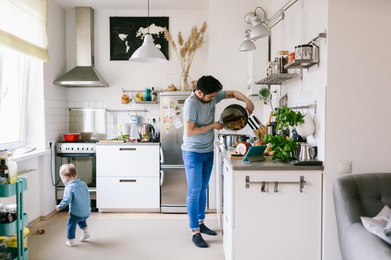 Picture shows a kitchen with different appliances and a man cooking on a stove and a child playing near him.